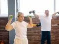 Mujer de 50 años tonifica sus brazos con mancuernas verdes. Un hombre a su lado también entrena.