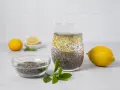 Vaso con agua de chia, rodaja de limón y semillas de chía en un bol. Limones y hojas verdes completan la escena.