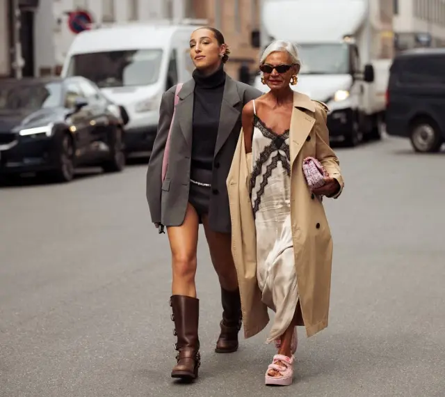 Dos mujeres pasean por la calle. Una lleva un vestido lencero de invierno con un abrigo y la otra un traje gris.