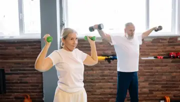 Mujer de 50 años tonifica sus brazos con mancuernas verdes. Un hombre a su lado también entrena.