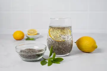Vaso con agua de chia, rodaja de limón y semillas de chía en un bol. Limones y hojas verdes completan la escena.