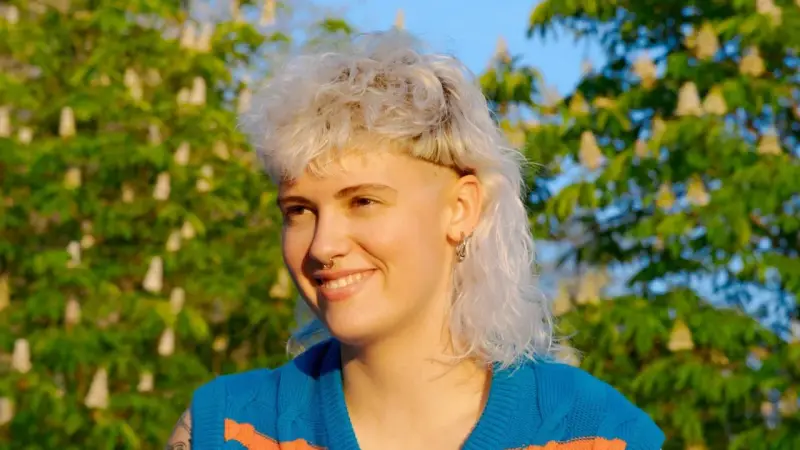 Persona sonriente con un moderno mullet rubio, aretes y un suéter azul con rayas naranjas, frente a un árbol en flor.